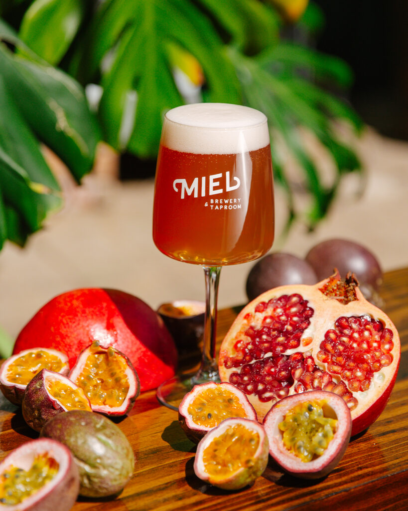 Miel fruited Frutsecco beer in a stemmed glass with a bright foam head, styled with fresh passion fruit and pomegranate in a tropical setting.