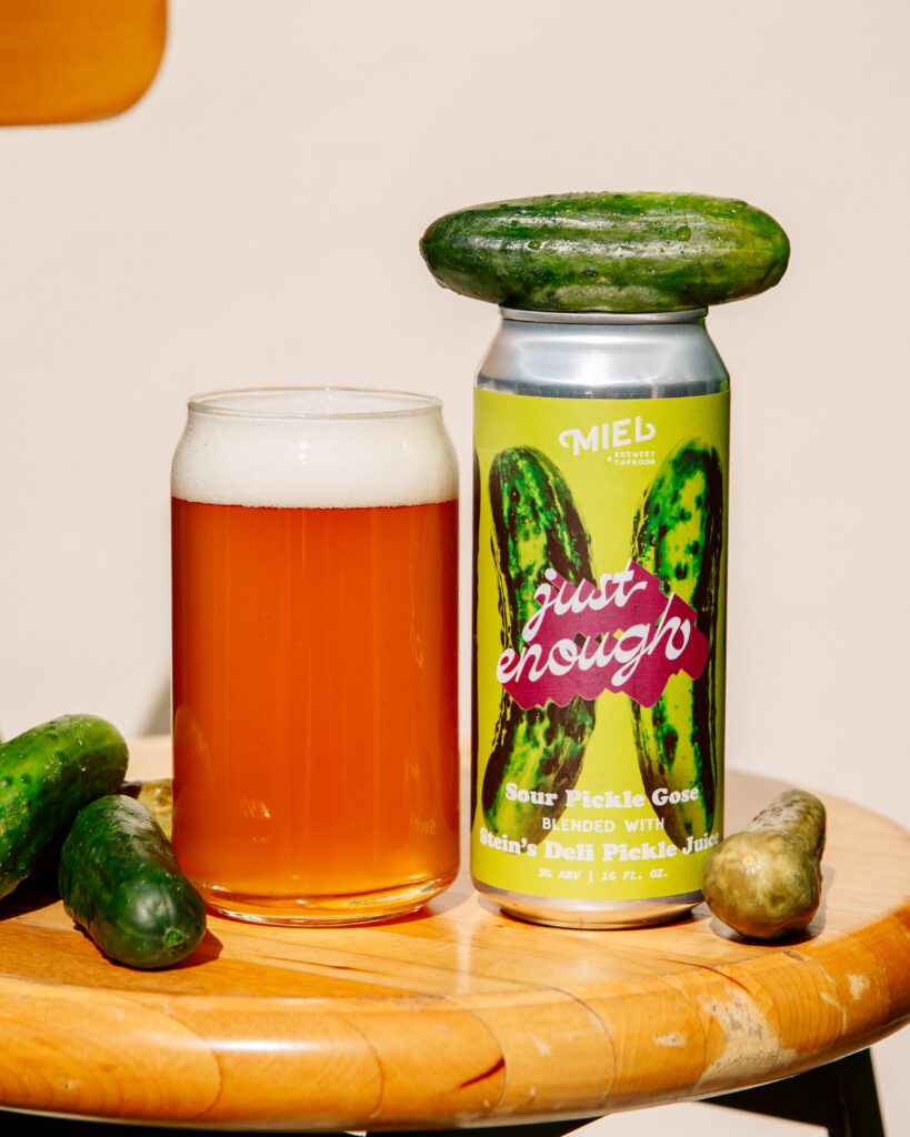 Miel Just Enough sour pickle gose with a foamy pour and playful pickle styling, featuring the can and fresh pickles on a wooden surface.