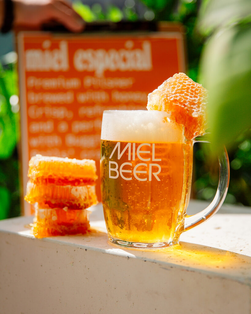 Glass mug of golden beer labeled “Miel Beer,” topped with foam and a piece of honeycomb, with stacked honeycomb beside it.