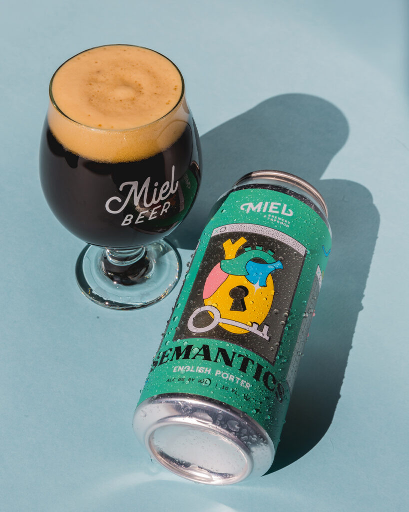 Tulip glass of dark Miel beer with a creamy foam head next to a chilled can of “Semantics” English Porter on a blue surface.